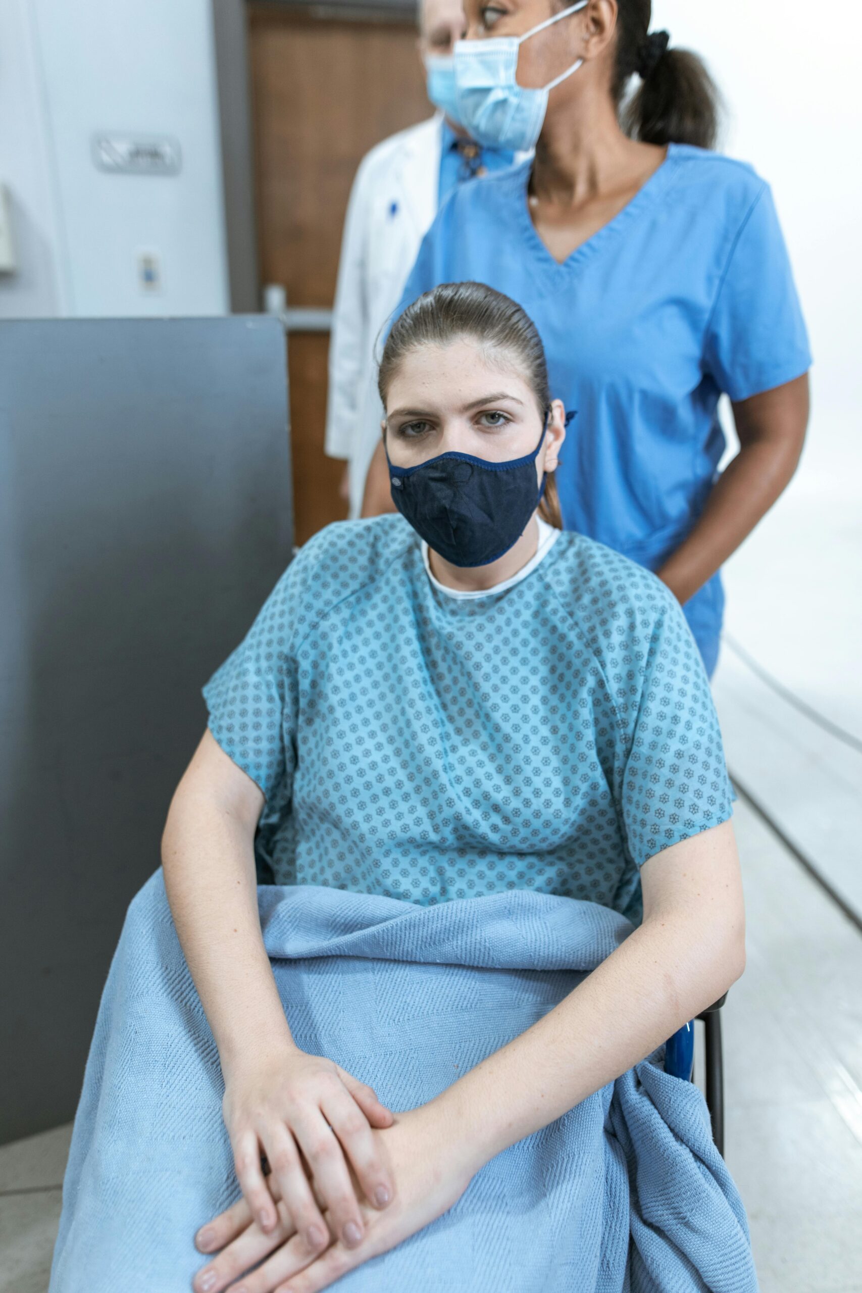 A nurse wearing scrubs assists a masked patient in a wheelchair, highlighting healthcare care.